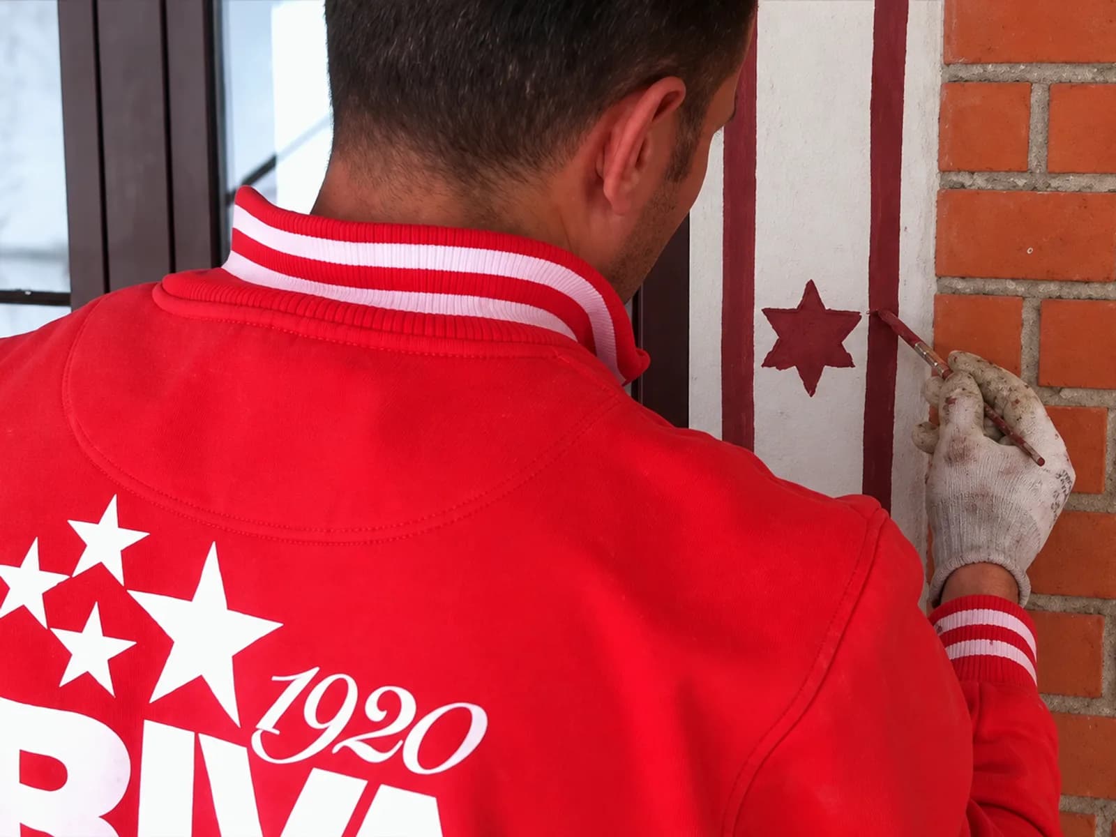 A RIVA worker, seen from behind in the firm’s red jacket, retouching a painted star on a pilaster beside a brick wall
